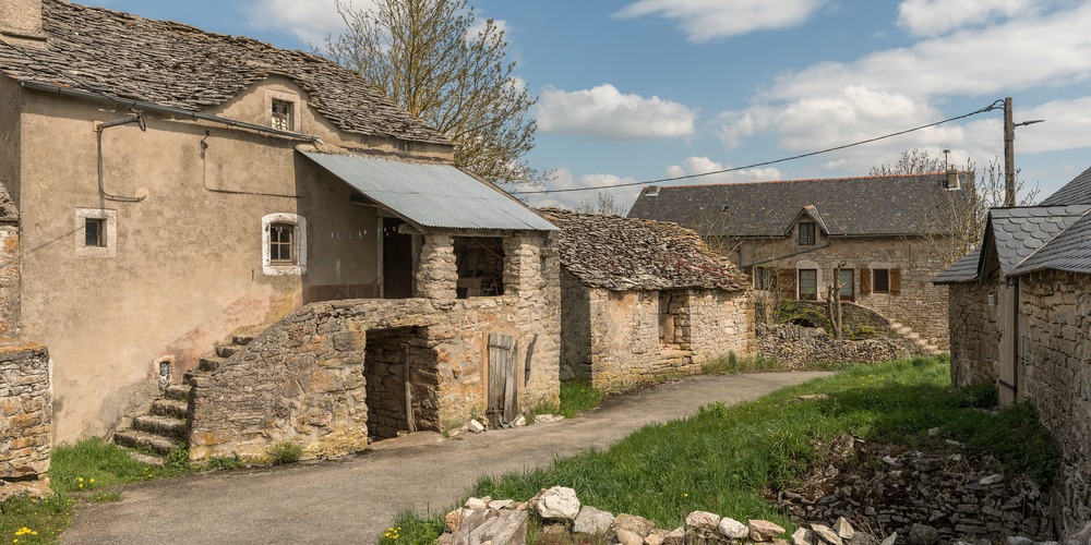 Hameau aux maisons de pierre et aux toits de lauzes, avec un escalier extérieur, un auvent de tôle et un chemin bordé d'herbe