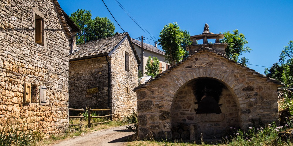 Ruelle de hameau en pierre avec un four à pain couvert de lauzes, des maisons aux volets de bois et un chemin de terre